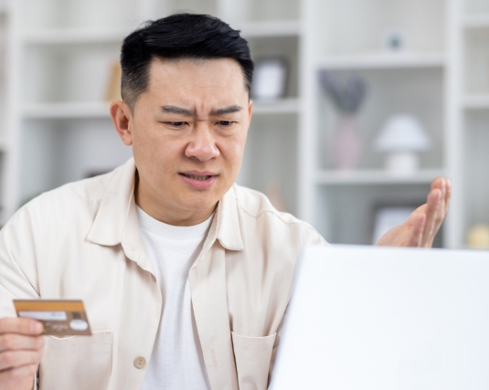 Asian man sitting at home at table holding bank credit card, rejected money transfer, negative account balance, upset and disappointed.