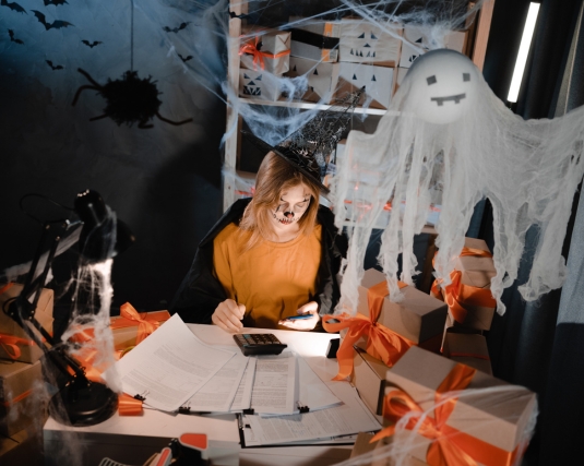 A woman in Halloween makeup and a witch costume counts finances on a calculator.