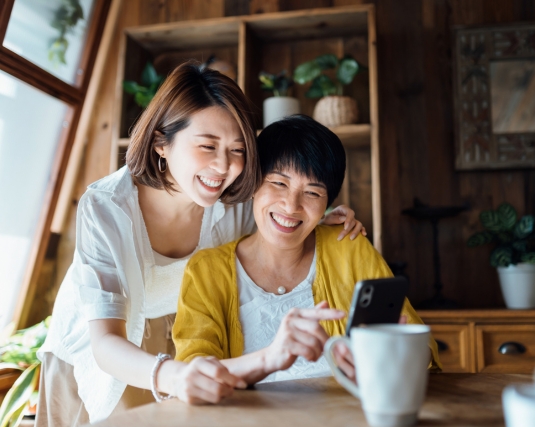 Affectionate Asian senior mother and daughter using smartphone together at home, smiling joyfully, enjoying mother and daughter bonding time.