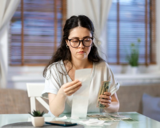 Portrait of pensive caucasian young woman accounting cash and paper receipt.