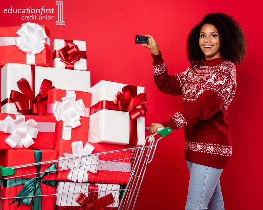 A woman holding up a credit card while pushing a shopping cart full of wrapped gifts.